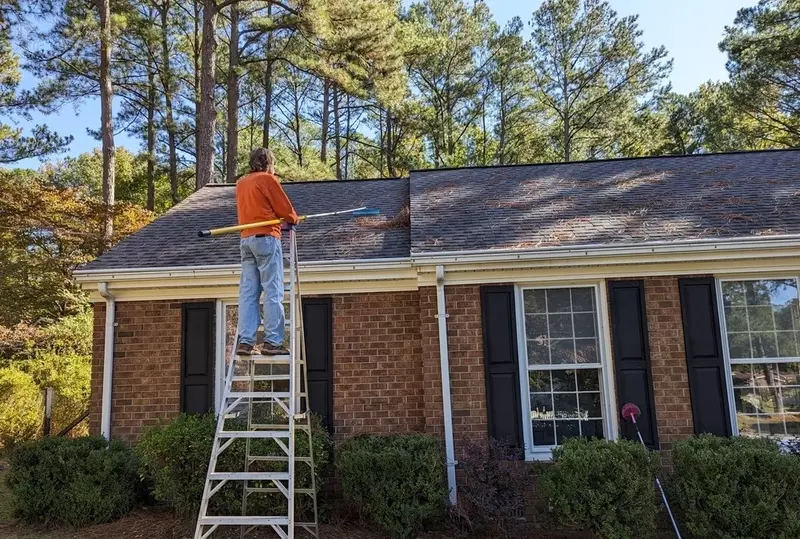 man raking pine needles off roof of North Carolina home