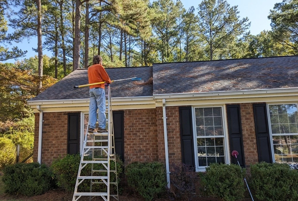 man raking pine needles off roof of North Carolina home